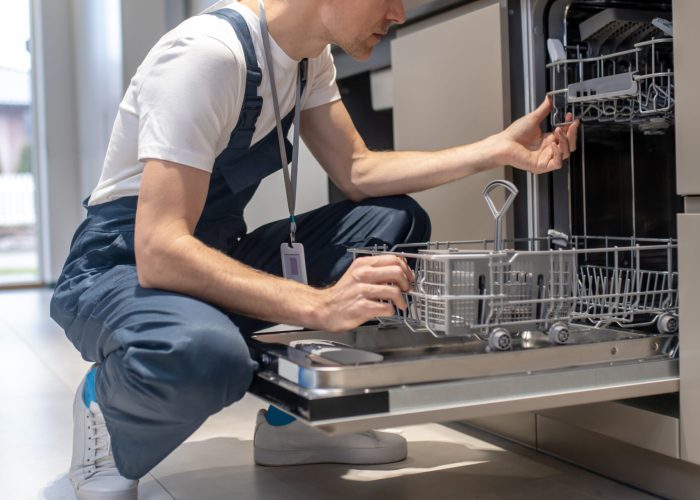 Diagnostics of equipment. Profile of serious man in dark overalls with badge crouching attentively looking at open dishwasher in kitchen at home during day